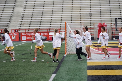A group of female athletes wearing white tops and yellow skirts are on a lacrosse field. Some are holding lacrosse sticks and running or walking across the field toward a goalpost. The setting appears to be a sports stadium with empty bleachers in the background.