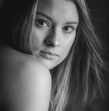 A close-up black and white portrait of a woman with long hair gazing directly at the camera. The light subtly highlights her facial features, especially her eyes and lips, contributing to an intimate and contemplative mood.