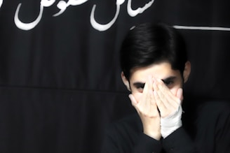 A person with short dark hair covers their mouth with their hands, while one hand is bandaged. The background is dark with some white cursive text partially visible.