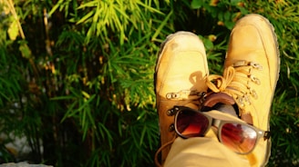 A pair of brown hiking boots resting near lush green foliage, with a pair of sunglasses placed on one of the boots suggesting relaxation in nature.