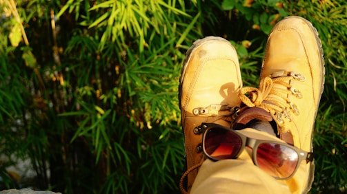 A pair of brown hiking boots resting near lush green foliage, with a pair of sunglasses placed on one of the boots suggesting relaxation in nature.