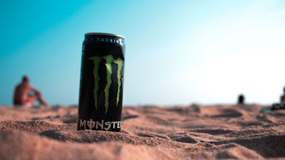 A can of energy drink is placed on sandy terrain with a blurred background of a beach and ocean. The sky appears clear with a light blue hue. The focus is on the can, which is prominent in the foreground, while people in the background are out of focus.