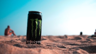 A can of energy drink is placed on sandy terrain with a blurred background of a beach and ocean. The sky appears clear with a light blue hue. The focus is on the can, which is prominent in the foreground, while people in the background are out of focus.