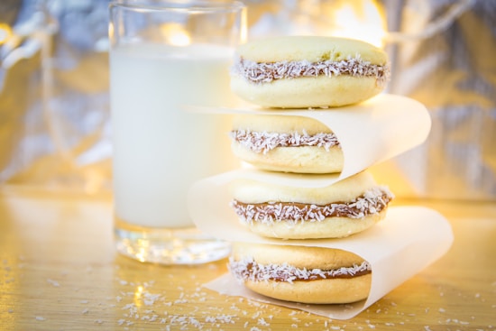 Three alfajores are stacked on a wooden surface, each filled with chocolate and coconut flakes along the edges. A glass of milk is placed behind them, partially out of focus. The background is softly illuminated, creating a cozy atmosphere.