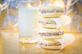 Three alfajores are stacked on a wooden surface, each filled with chocolate and coconut flakes along the edges. A glass of milk is placed behind them, partially out of focus. The background is softly illuminated, creating a cozy atmosphere.