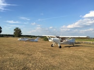 A grassy field under a clear blue sky with scattered clouds. Two small aircraft are parked on the field; one is a single-engine propeller plane, and the other is a glider. The field is bordered by a wooden fence, and there are trees and shrubs in the background.