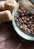 A glass container is filled with roasted coffee beans, placed on a textured brown surface alongside several wine corks. The mixture of light and shadow creates a cozy and rustic atmosphere.
