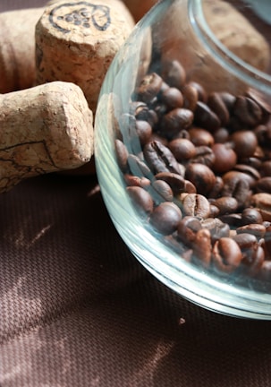 A glass container is filled with roasted coffee beans, placed on a textured brown surface alongside several wine corks. The mixture of light and shadow creates a cozy and rustic atmosphere.