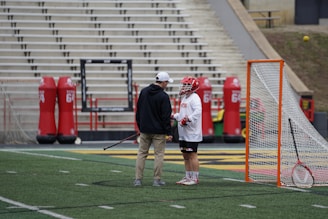 A lacrosse player wearing a helmet and gloves stands on the field next to a coach or trainer, both engaged in conversation. The player is holding a lacrosse stick. Nearby, there is a lacrosse goal and red practice equipment on the field. The stadium seating in the background is empty.