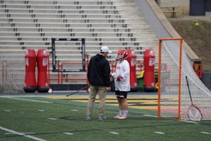 A lacrosse player wearing a helmet and gloves stands on the field next to a coach or trainer, both engaged in conversation. The player is holding a lacrosse stick. Nearby, there is a lacrosse goal and red practice equipment on the field. The stadium seating in the background is empty.