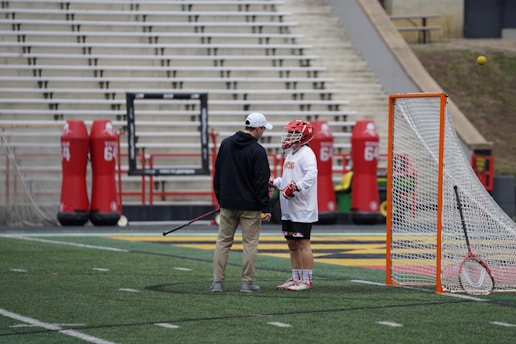 A lacrosse player wearing a helmet and gloves stands on the field next to a coach or trainer, both engaged in conversation. The player is holding a lacrosse stick. Nearby, there is a lacrosse goal and red practice equipment on the field. The stadium seating in the background is empty.