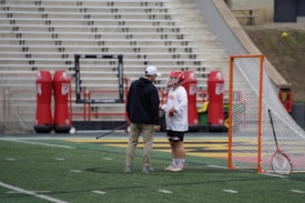 A lacrosse player wearing a helmet and gloves stands on the field next to a coach or trainer, both engaged in conversation. The player is holding a lacrosse stick. Nearby, there is a lacrosse goal and red practice equipment on the field. The stadium seating in the background is empty.