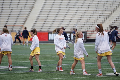 A group of young women wearing athletic gear are on a sports field. They are dressed in white jerseys with yellow shorts, featuring a distinctive design at the hem. The field has a green turf surface and the background shows empty bleachers. Some additional figures are visible in the distance, possibly referees or other players.