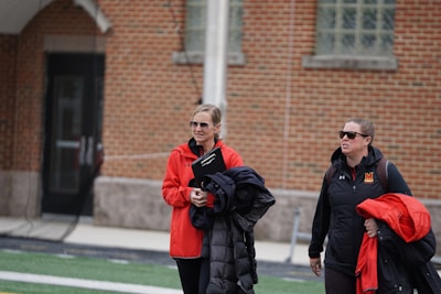 Two individuals wearing sports jackets and sunglasses are walking together outdoors, one holding a clipboard and a coat. They are near a brick building with arched windows and a door visible in the background.