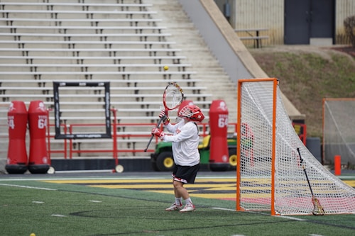 A lacrosse player in protective gear stands in front of a goal net, attempting to catch a ball with a lacrosse stick. The background features empty stadium bleachers and red tackling dummies.