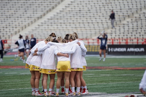 A group of athletes wearing white jerseys and yellow shorts stand in a tight huddle on a sports field. The stadium seats behind them are mostly empty, and a few other people can be seen in the background, some in motion.