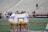 A group of athletes wearing white jerseys and yellow shorts stand in a tight huddle on a sports field. The stadium seats behind them are mostly empty, and a few other people can be seen in the background, some in motion.