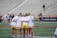 A group of athletes wearing white jerseys and yellow shorts stand in a tight huddle on a sports field. The stadium seats behind them are mostly empty, and a few other people can be seen in the background, some in motion.