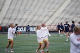 A group of women wearing sports uniforms are on a field, possibly preparing for a lacrosse game. They are engaged in light activity, with some in the background playing with lacrosse sticks. The seating area of the stadium is visible, suggesting this is a practice session or warm-up before a game.