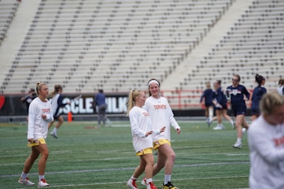 A group of women wearing sports uniforms are on a field, possibly preparing for a lacrosse game. They are engaged in light activity, with some in the background playing with lacrosse sticks. The seating area of the stadium is visible, suggesting this is a practice session or warm-up before a game.