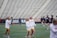 A group of women wearing sports uniforms are on a field, possibly preparing for a lacrosse game. They are engaged in light activity, with some in the background playing with lacrosse sticks. The seating area of the stadium is visible, suggesting this is a practice session or warm-up before a game.