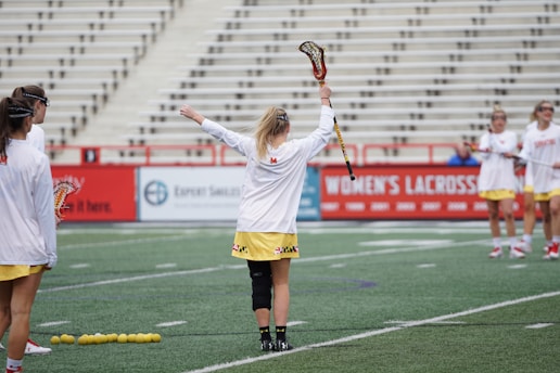 A group of women wearing sports uniforms practicing on a lacrosse field. They are holding lacrosse sticks and some yellow balls are gathered on the ground. The background shows a largely empty stadium with rows of seating.