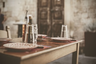 A rustic dining table is set with empty plates, glasses turned upside down, and bottles of olive oil and vinegar. The background features a weathered stone wall and a wooden door, suggesting a cozy, old-world ambiance.