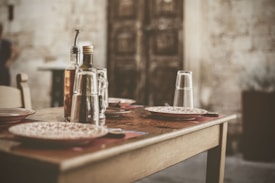 A rustic dining table is set with empty plates, glasses turned upside down, and bottles of olive oil and vinegar. The background features a weathered stone wall and a wooden door, suggesting a cozy, old-world ambiance.