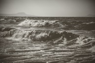 Waves crash energetically against each other, showing the dynamic movement of the ocean. The sea is turbulent, with frothy whitecaps visible on the surface. In the background, there are faint silhouettes of distant landmasses under an overcast sky.
