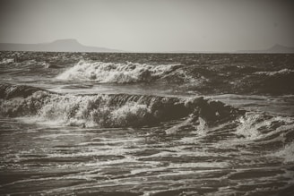 Waves crash energetically against each other, showing the dynamic movement of the ocean. The sea is turbulent, with frothy whitecaps visible on the surface. In the background, there are faint silhouettes of distant landmasses under an overcast sky.