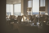 A dimly lit, elegantly set restaurant table near large windows. Napkins are folded in a standing position, and the table is arranged with upside-down wine glasses and silverware. The ambiance suggests a calm and sophisticated dining environment.