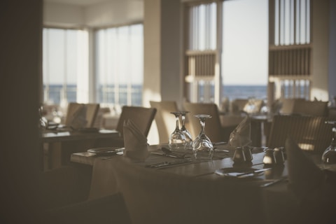 A dimly lit, elegantly set restaurant table near large windows. Napkins are folded in a standing position, and the table is arranged with upside-down wine glasses and silverware. The ambiance suggests a calm and sophisticated dining environment.