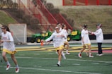 A group of women wearing sports gear, including helmets and lacrosse sticks, are engaging in a sports practice on a field with marked lines. They are dressed in white shirts and yellow shorts, with some wearing visors or protective eyewear. In the background, there is a tractor and a net goal, along with red metal railings and brown shrubbery.