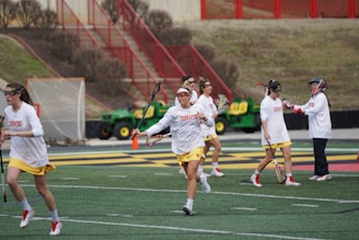 A group of women wearing sports gear, including helmets and lacrosse sticks, are engaging in a sports practice on a field with marked lines. They are dressed in white shirts and yellow shorts, with some wearing visors or protective eyewear. In the background, there is a tractor and a net goal, along with red metal railings and brown shrubbery.