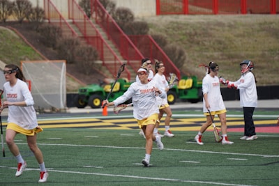 A group of women wearing sports gear, including helmets and lacrosse sticks, are engaging in a sports practice on a field with marked lines. They are dressed in white shirts and yellow shorts, with some wearing visors or protective eyewear. In the background, there is a tractor and a net goal, along with red metal railings and brown shrubbery.