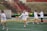 A group of women wearing sports gear, including helmets and lacrosse sticks, are engaging in a sports practice on a field with marked lines. They are dressed in white shirts and yellow shorts, with some wearing visors or protective eyewear. In the background, there is a tractor and a net goal, along with red metal railings and brown shrubbery.