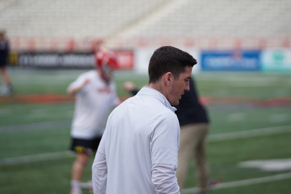 A person with short dark hair is wearing a white jacket and standing near a sports field. In the background, another person wearing a red helmet and white sports attire appears to be engaged in some form of athletic activity. The setting is a stadium with blurred advertisements on the walls.