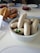 A dining table setup features a large white bowl filled with white sausages garnished with a sprig of parsley. Nearby, a basket contains pretzels and bread rolls. A few red coffee cups and saucers are placed on the table along with plates, some containing more sausages.