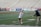 A lacrosse player in a red helmet and white jersey is being coached by another person wearing a hat and dark sweatshirt on a sports field. The player is reaching out with one hand while holding a lacrosse stick in the other, and there are bleachers in the background.