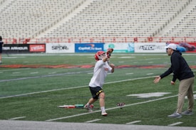 A lacrosse player in a red helmet and white jersey is being coached by another person wearing a hat and dark sweatshirt on a sports field. The player is reaching out with one hand while holding a lacrosse stick in the other, and there are bleachers in the background.