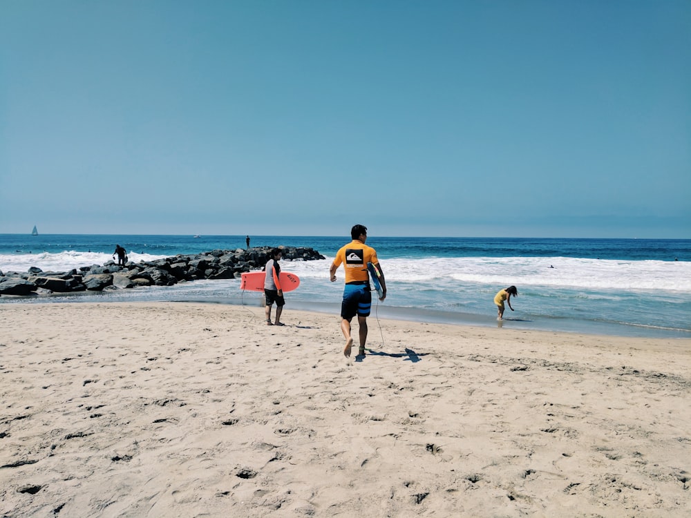 two person walking on beach shore holding bodyboards