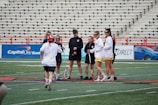 A group of women is standing together on a sports field, holding lacrosse sticks. They are wearing sports jerseys and shorts, some in red and white colors. The background has empty bleachers and advertisement banners visible.