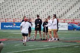 A group of women is standing together on a sports field, holding lacrosse sticks. They are wearing sports jerseys and shorts, some in red and white colors. The background has empty bleachers and advertisement banners visible.