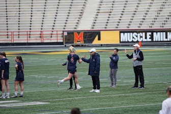 Several people are on a sports field with an empty bleacher in the background. One person is high-fiving a coach, while others stand nearby. They are wearing athletic clothing, hinting at a team sport environment. A sign with the text 'Muscle Milk' is visible in the background.