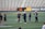 Several people are on a sports field with an empty bleacher in the background. One person is high-fiving a coach, while others stand nearby. They are wearing athletic clothing, hinting at a team sport environment. A sign with the text 'Muscle Milk' is visible in the background.