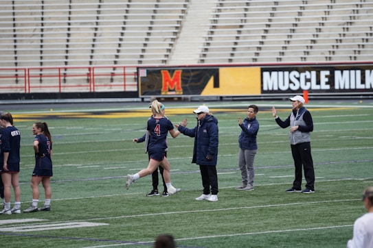 Several people are on a sports field with an empty bleacher in the background. One person is high-fiving a coach, while others stand nearby. They are wearing athletic clothing, hinting at a team sport environment. A sign with the text 'Muscle Milk' is visible in the background.
