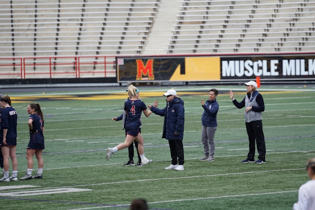 Several people are on a sports field with an empty bleacher in the background. One person is high-fiving a coach, while others stand nearby. They are wearing athletic clothing, hinting at a team sport environment. A sign with the text 'Muscle Milk' is visible in the background.