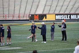 Several people are on a sports field with an empty bleacher in the background. One person is high-fiving a coach, while others stand nearby. They are wearing athletic clothing, hinting at a team sport environment. A sign with the text 'Muscle Milk' is visible in the background.