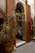 An ornate doorway framed by intricate wooden carvings and detailed mosaic tiles. The scene includes a potted plant with long, thin leaves and a traditional Moroccan lantern placed on a geometric patterned tile floor.