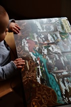 A child is seated and looking at a colorful illustrated book depicting a wintry forest scene with snow, trees, and a flowing river. The illustration includes various animals, such as a red bird and possibly other small creatures. The child is wearing a gray long-sleeve shirt and has their hands resting on the book. The word in Cyrillic at the top edge likely refers to a month or season.